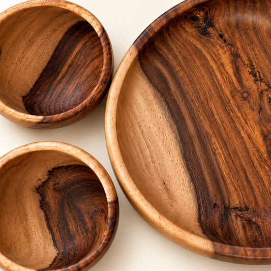 Three handcrafted wooden serving dishes with rich natural wood grain patterns, including two bowls and one large platter, arranged on a light cream background.